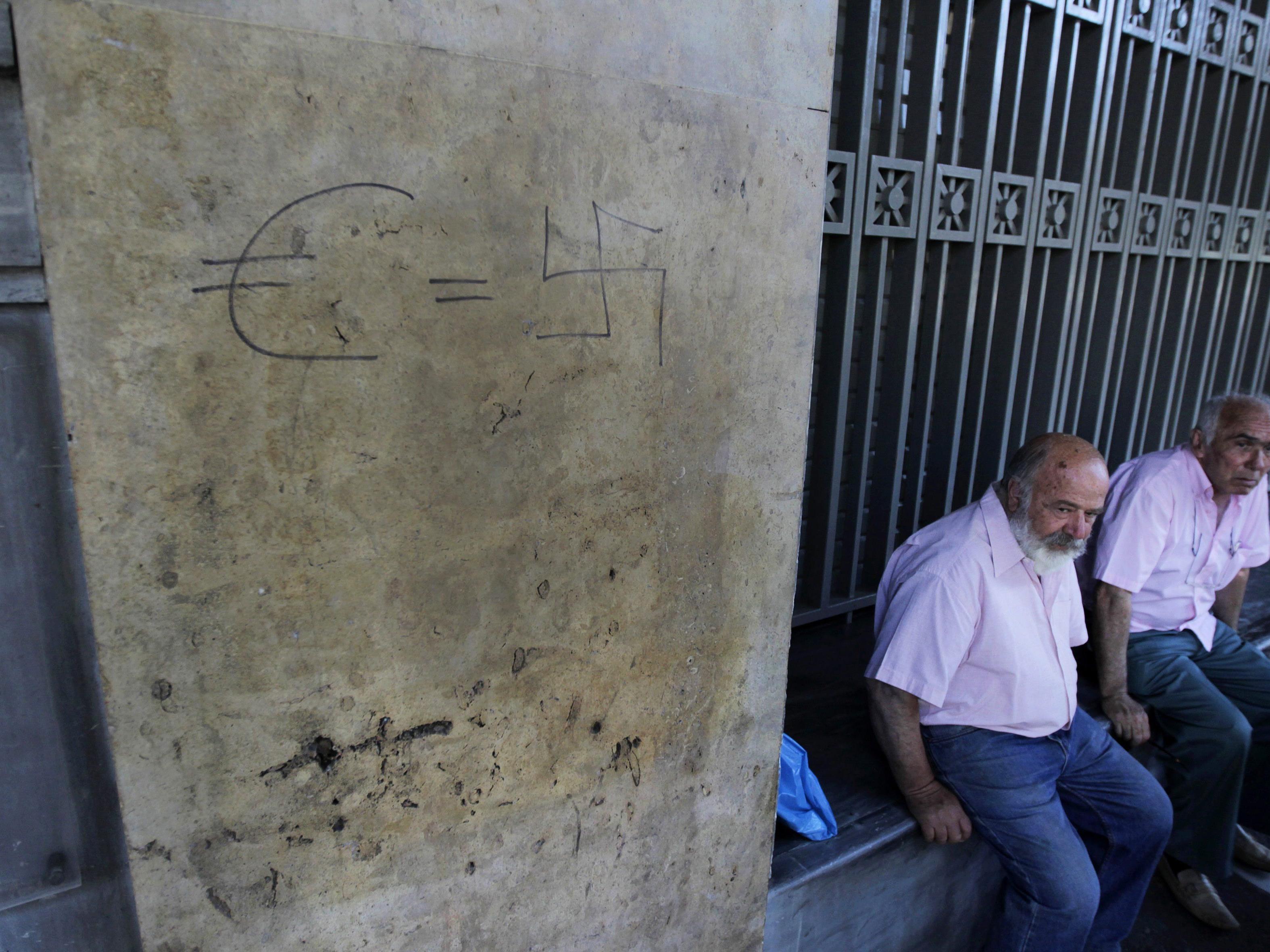 Zwei Pensionisten vor einer geschlossenen Bank. Ein Graffiti an der Wand setzt den Euro mit dem Nationalsozialismus gleich.